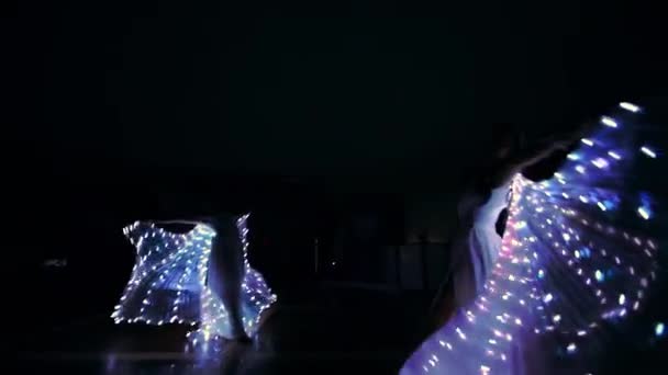 Las chicas realizan un baile con alas led. Las luces led brillan en la oscuridad. Las chicas jóvenes giran en torno a sí mismas y bailan en la oscuridad. — Vídeos de Stock