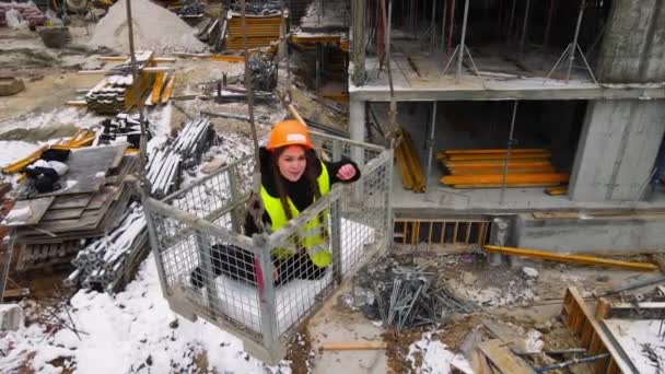 Žena na staveništi v zimě, zvednutá jeřábem. Žena mluví text do kamery, zatímco je v košíku nad staveništěm. — Stock video