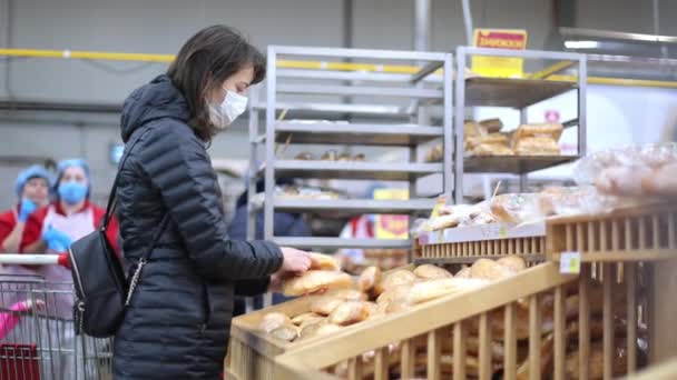 Egy nő a szupermarketben a karantén alatt védőmaszkban kenyeret vesz. Vásárolni egy szupermarketben. Pékipari termékeket vásárló nő egy szupermarketben pékségben — Stock videók