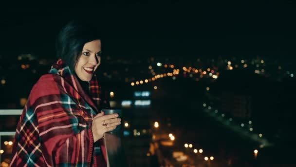 Mujer feliz en la terraza de la casa tomando té caliente en el fondo de la ciudad de la noche. La chica está envuelta en una manta caliente. Concepto de mujer feliz y exitosa. — Vídeos de Stock