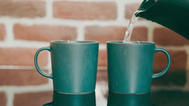 Preparando té para dos personas. Verter agua caliente en tazas de té verde de pie sobre una superficie de amor espejada sobre un fondo de pared de ladrillo rojo. Té de la mañana para dos temprano en la mañana. — Vídeos de Stock