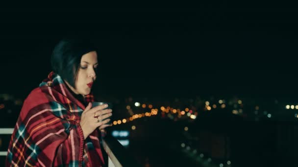 Mujer feliz en la terraza de la casa tomando té caliente en el fondo de la ciudad de la noche. La chica está envuelta en una manta caliente. Concepto de mujer feliz y exitosa. — Vídeos de Stock