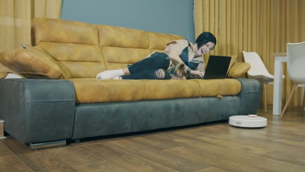 Een vrouw die op een bank ligt en aan een laptop werkt, en een robot stofzuiger stofzuigen automatisch de kamer. Automatische schoonmaak. Vrije tijd om met uw huisdier te werken. — Stockvideo