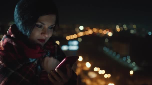 Donna usa un telefono cellulare di notte e sorride emotivamente e reagisce a ciò che vede, sullo sfondo della città di notte. La luce del telefono cade sul volto delle donne. — Video Stock
