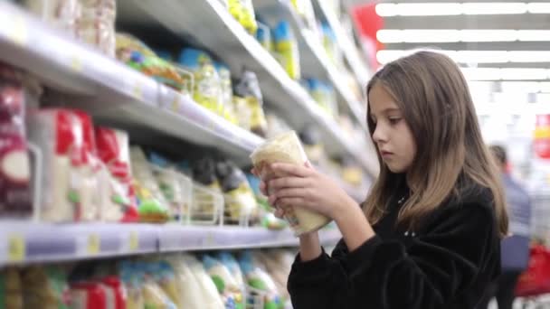 Девочка выбирает товары в супермаркете. Ребенок забирает товар на полке супермаркета и осматривает его. Концепция детских покупок на продуктовом рынке — стоковое видео