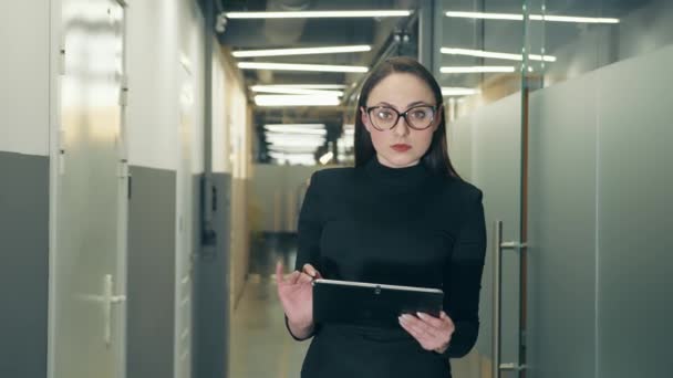 Retrato de una exitosa mujer de negocios con gafas y ropa negra de apariencia europea parada en el pasillo y sosteniendo una tableta. Un profesional trabaja en un dispositivo táctil — Vídeos de Stock