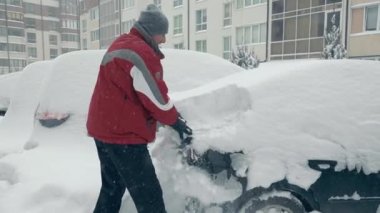 Ukrayna Lviv 10.02.2021. Kar yağışı ve fırtınalar. Bir adam karla kaplı bir arabayı temizler. Şiddetli kar yağışı. Sırt çantalı ve fırçalı sürücü park yerinde duran arabadan karı temizliyor.