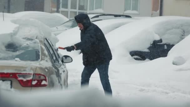 Dérives de neige et tempêtes. Un homme nettoie une voiture enneigée. De fortes chutes de neige. Le conducteur dans un sac à dos avec une brosse efface la neige de la voiture debout sur le parking — Video