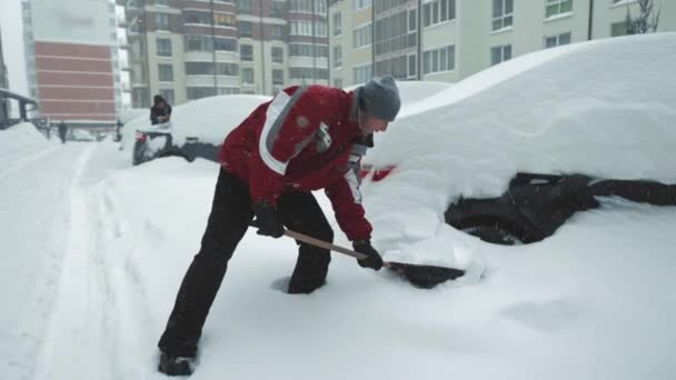 Egy férfi megtisztítja a kocsiját a hótól. Eltakarítani a havat a parkolóból egy lapáttal.. — Stock videók