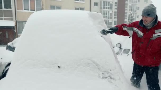 Χιόνι παρασύρεται και καταιγίδες. Ένας άντρας καθαρίζει ένα χιονισμένο αυτοκίνητο. Βαριά χιονόπτωση. Ο οδηγός σε σακίδιο με βούρτσα καθαρίζει το χιόνι από το αυτοκίνητο που στέκεται στο πάρκινγκ — Αρχείο Βίντεο