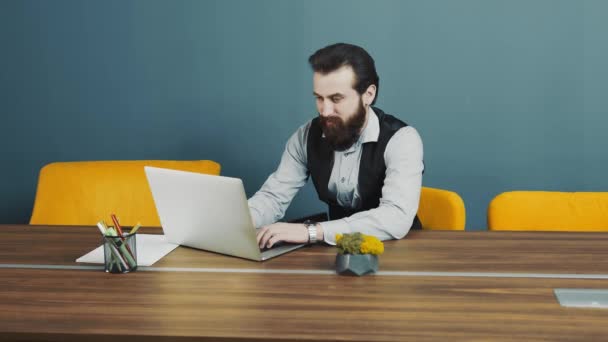 Молодий бородатий чоловік програмує кавказьку зовнішність, сидячи і працюючи на ноутбуку. Стреляю через бокал офиса. Обличчя від скла проходить крізь раму в русі.. — стокове відео