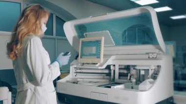 Laboratuvardaki kadın kullanıyor. Mobil cihaz biyokimyasal kan analizöründen bilgi alır. Tıbbi maskeli bir laboratuvar çalışanı cep telefonuna bakıyor.