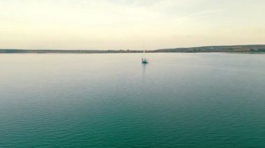Gün batımında denizde bir yatın üstünde hava manzarası. Güneş ve bulutlar suya yansıyor. Yolculuğa çıkan bir yat..
