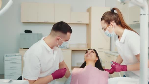 O dentista junto com o assistente examinam o paciente na clínica odontológica. O médico pergunta ao paciente sobre problemas dentários. Comunicação entre médico e paciente — Vídeo de Stock