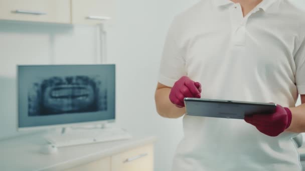 Hände eines Arztes, der die Touchtasten eines Tablets drückt. Nahaufnahme einer Arzthand mit einem Tablet. Im Hintergrund ein Röntgenbild eines Kiefers auf einem Computermonitor. — Stockvideo