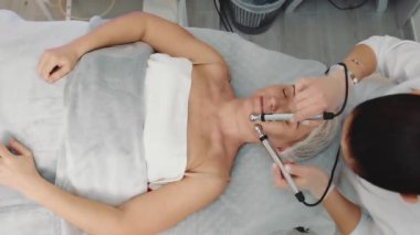 Microcurrent therapy top view. A cosmetologist conducts microcurrent therapy for an elderly woman. The procedure of rejuvenation and restoration of old skin