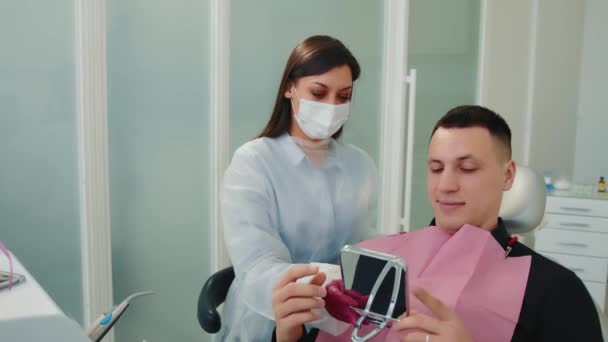 El dentista le da un espejo al paciente para evaluar el trabajo del dentista. El paciente mira sus dientes en el espejo — Vídeos de Stock