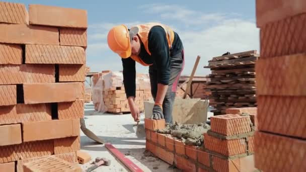 Közelkép egy férfiról, aki téglaházat épít. Piros téglákat fektetni egy építkezésen egy napsütéses napon. Lakásépítés közelről. — Stock videók