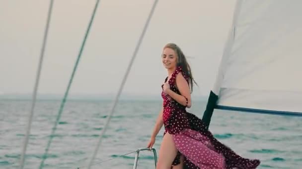 Menina bonita jovem em um iate à noite ao pôr-do-sol. Uma viagem de iate no alto mar. Jovem feliz sorri e levanta a mão para cima da colina. Forte vento levanta o vestido das meninas. — Vídeo de Stock