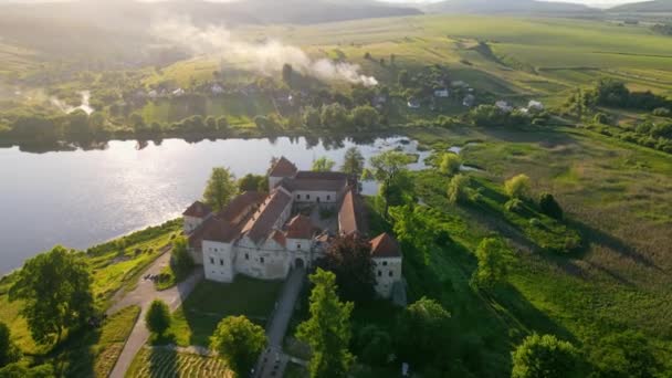 Hrad u jezera z ptačí perspektivy při západu slunce. Starobylý hrad v malebném místě v obci, v blízkosti jezera a na slunci. Bílý kouř se šíří po vesnici — Stock video