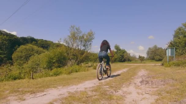 Vista da parte de trás de uma menina andando de bicicleta na estrada. Andar de bicicleta numa estrada de campo. Jovem mulher pedala uma bicicleta — Vídeo de Stock