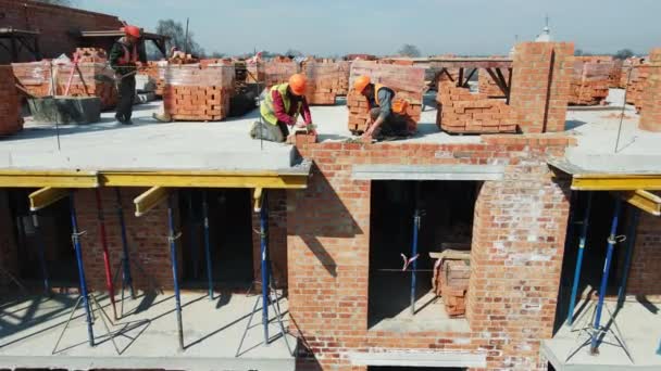 Egy vörös téglákat fektető ház építésén dolgozó munkások. Az építők téglafalat építenek egy toronyházból.. — Stock videók