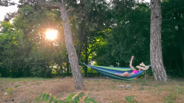 Szczęśliwa młoda kobieta leżąca w hamaku w parku o zachodzie słońca z kapeluszem na twarzy. Wspaniały odpoczynek w parku na hamaku. dziewica w stroju kąpielowym leży w hamaku o zachodzie słońca. — Wideo stockowe