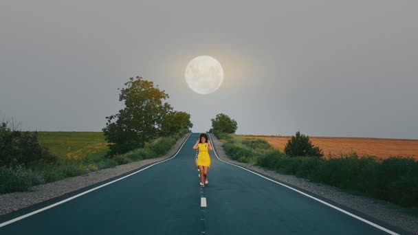 穿着黄色连衣裙在路上跑的女人。大月亮照亮了道路.快乐的女人把她的手抬起来. — 图库视频影像