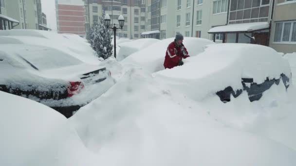 Сніг дрейфує і шторми. Чоловік чистить сніговий автомобіль. Сильний снігопад. Водій в рюкзаку з пензлем розчищає сніг від машини, що стоїть на парковці — стокове відео