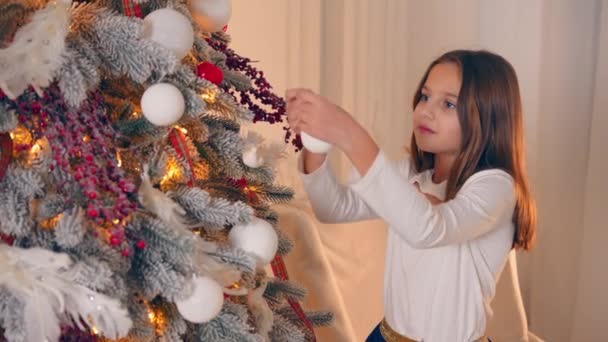 Petite fille décore le sapin de Noël avec des décorations. Préparatifs pour Noël et Nouvel An 2022 — Video