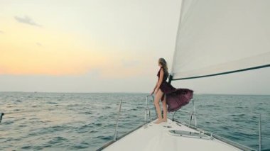 Gece elbisesi giymiş bir kız gün batımında yatta duruyor. İyi akşamlar tekne gezisi. Zarif elbiseli kadın rüzgârı kaldırıyor. Açık denizde bir yatta mutlu bir kadın.