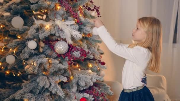 Liten flicka dekorerar julgranen och förbereder sig för jul och nyår. Ett litet barn sätter juldekorationer på julgranen. — Stockvideo