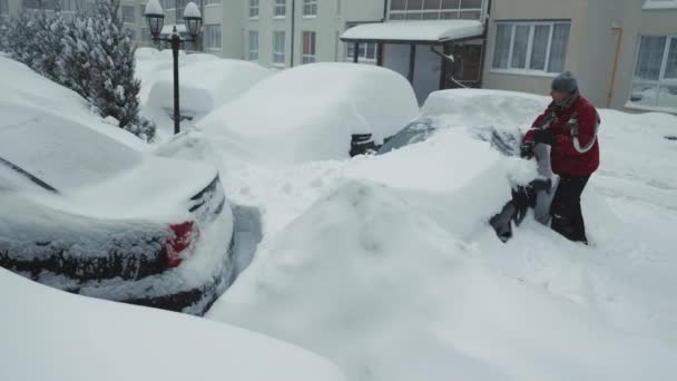 Sněhové závěje a bouře. Muž vyčistí zasněžené auto. Silný sníh. Řidič v batohu se štětcem čistí sníh z auta stojícího na parkovišti — Stock video