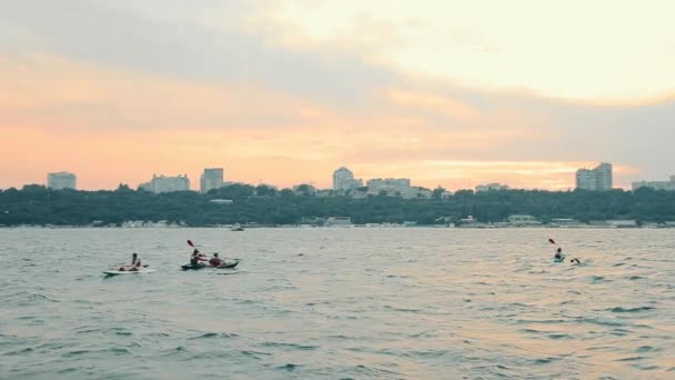 Emberek a tengerben úszni nedv tálcák és kenuk naplementekor a háttérben a város. — Stock videók