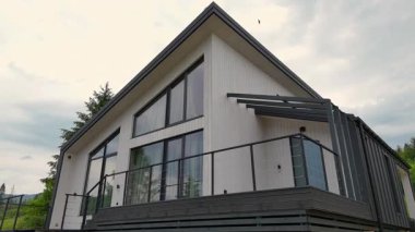 Minimalist modern kır evi, yüksek perdeli çatı, geniş pencere cephesi ve bulutlu bir gökyüzüne karşı geniş güverte..