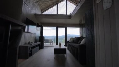 Geniş panoramik pencereli ve dağ manzaralı modern ahşap kulübe. Gri kanepesi ve dinlenmek için şöminesi olan rahat oturma odası..