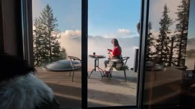 Sabahları terasta oturan ve kahve içen genç bir kadın cam kapının ardındaki sisli dağların güzel manzarasının tadını çıkarıyor..