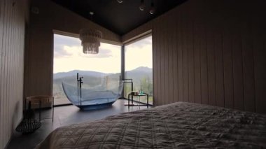 Modern lüks yatak odası pencerenin yanında şık mavi cam küvet, bulutlu bir günde rahat otel odasından güzel dağ manzarası..