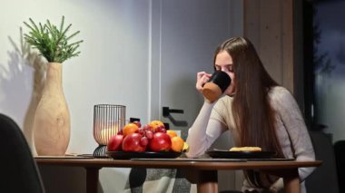 Uzun saçlı genç bir kadın yemek masasında siyah bardaktan çay içiyor. Evde sıcak bir akşam ve modern odada meyve tabağı ve kış süsüsüyle..