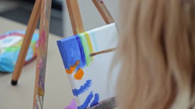 Küçük tuvale fırçayla çocuk eli boyama sehpasında, genç ressam mavi boyayla renkli ve soyut bir resim yaratıyor..