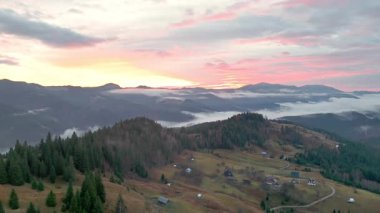 Pembe günbatımında vadide sis varken yeşil tepelerde dağınık evleri olan dağ köyünün hava panoramik manzarası..