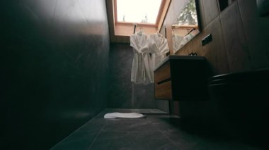 Modern banyo içi koyu taş duvarlar ve ahşap aksanlı, iki beyaz bornoz evdeki tavan penceresinin altında asılı..