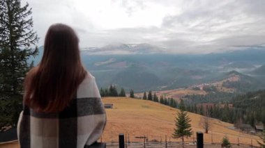Sisli tepelere bakan bir kadın, balkonda battaniyeye sarılı bir kız..