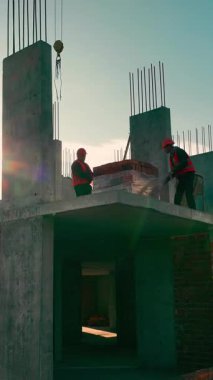 Bir tuğla ev inşa etmek. İnşaatçılar bir paleti tuğlalarla açıyorlar. İnşaat alanında çalışma süreci. İnşaat alanındaki inşaatçıların siluetleri. Güneş ışınları kameraya yansıyor..