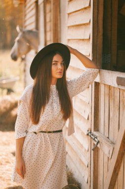 Güneşli bir günde çiftlikte poz veren şapkalı ve elbiseli genç bir kız.