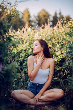 Kolsuz tişörtlü, kısa şortlu genç esmer çimenlerde otururken poz veriyor.