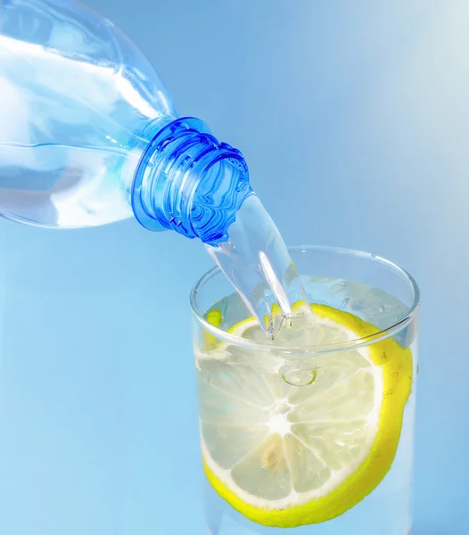 Gießen von Wasser aus der Flasche in Glas mit Zitrone auf blauem Hintergrund — Stockfoto