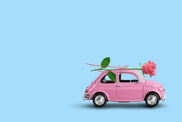 Pink toy car delivering pink rose flower on a blue background. Flowers delivery, women day, valentine day.