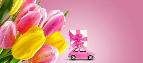 Pink retro car with gift box on a roof with tulip flowers on pink background. Greeting card, banner design template