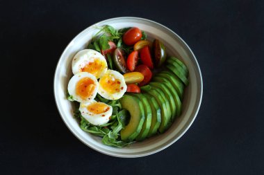 Akdeniz salatası, avokado ile haşlanmış yumurta, kremalı yumurta ve roket yatağında vişneli domates, EVO yağı, tuz ve limon suyu. Tek ve dengeli yemek. Siyah arka plan. Genel Görünüm.